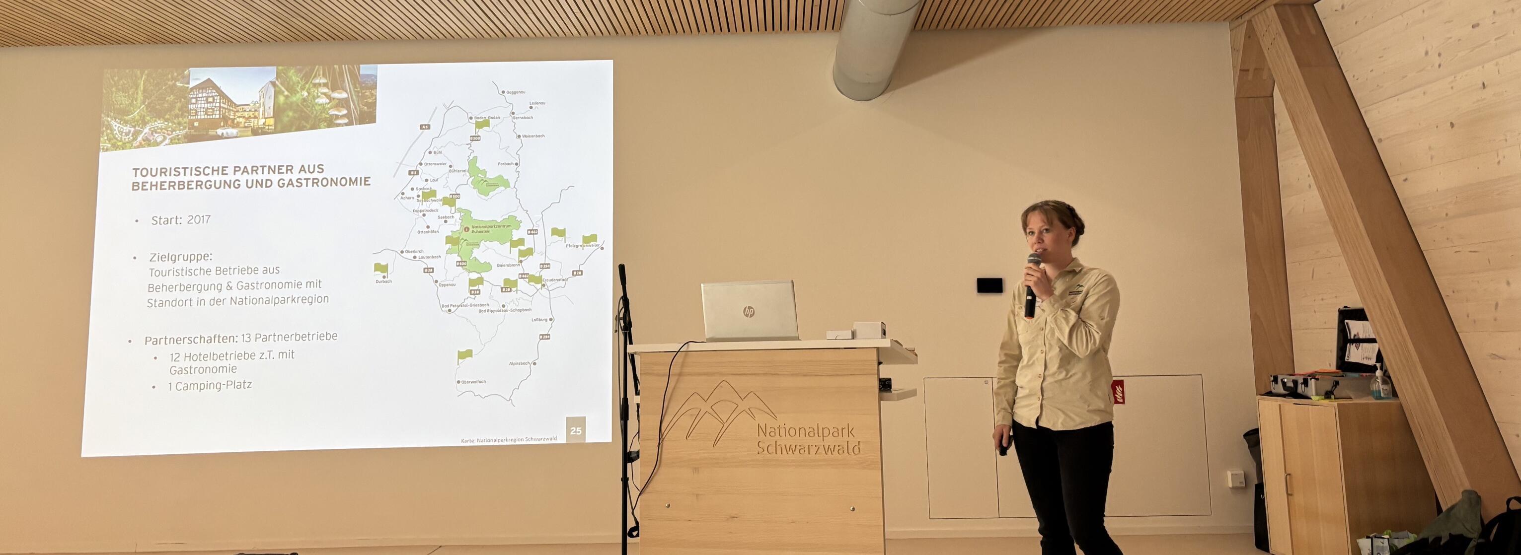 Eine Frau steht in einem Saal mit viel Holz vor neben einer Leinwand und einem Rednerpult. Foto: Natalie Schacht