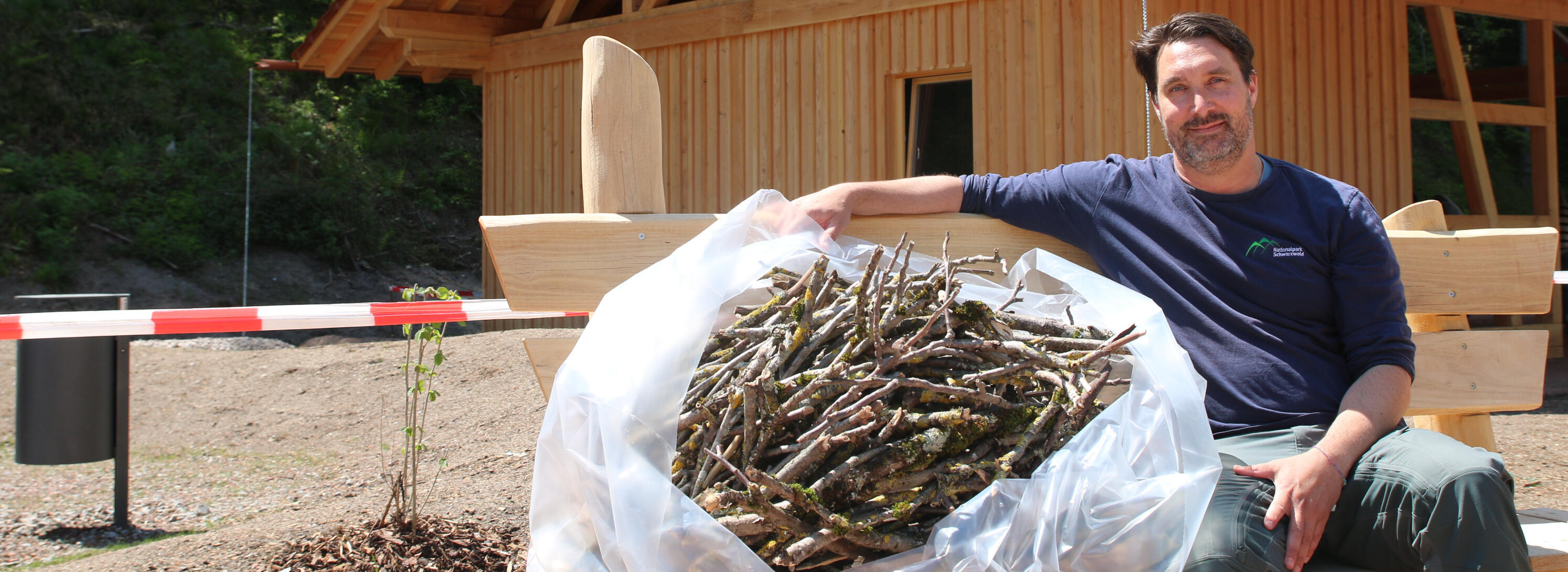 Ein Mann im blauen Tshirt sitzt neben einem Sack mit Stöcken auf einer Bank, im Hintergrund eine Holzhütte. Foto: Anne Kobarg (Nationalpark Schwarzwald)
