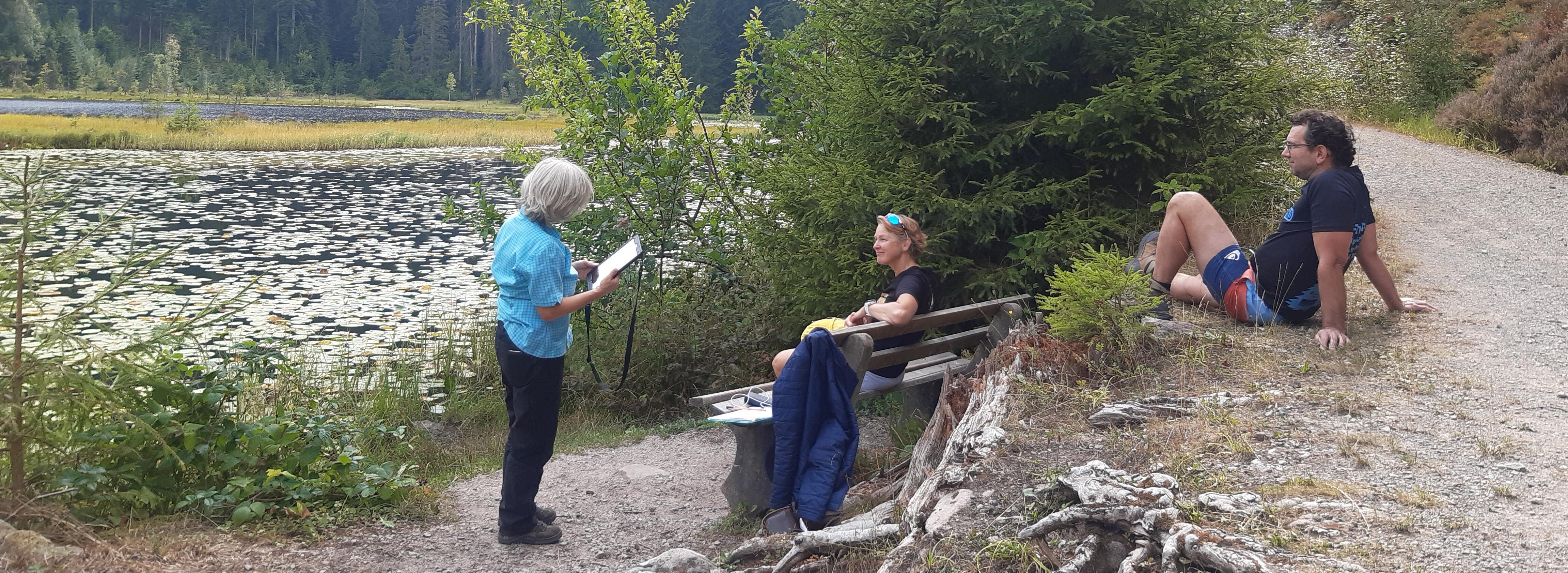 Am Seeufer führt eine Frau ein Interview mit einer Frau, die auf einer Bank sitzt. Ein Mann sitzt daneben auf einem Weg.