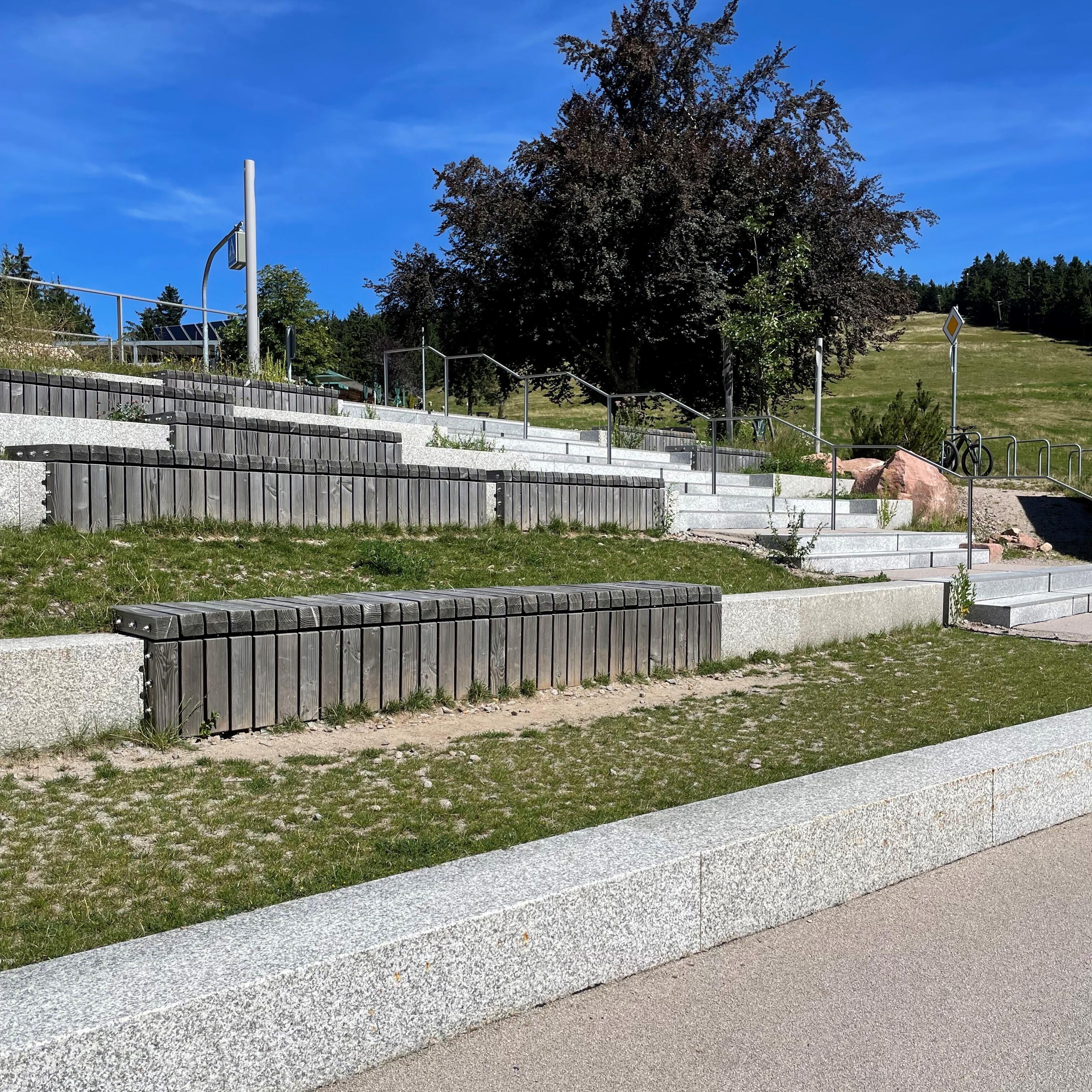 Gestufter Sitzbereich wie in Stadion neben Treppe, teilweise mit Holz belegt. Bodenbelag: Rasen und Sand.