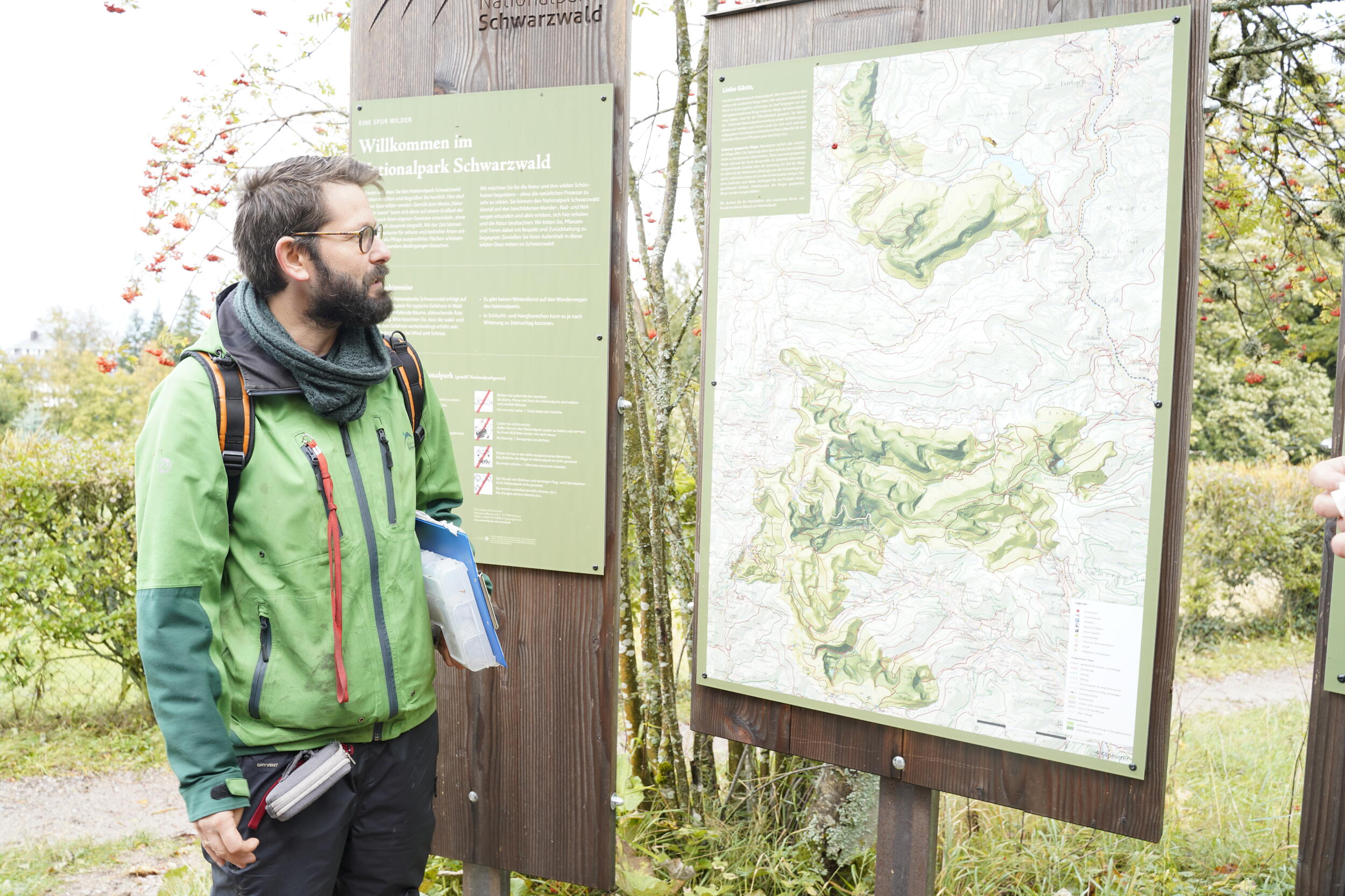 Ein Mann mit dunklen Haaren und Bart in grüner Nationalparkjacke steht vor einer Übersichtskarte im Nationalpark Schwarzwald. @ Judith Wildt Bastos