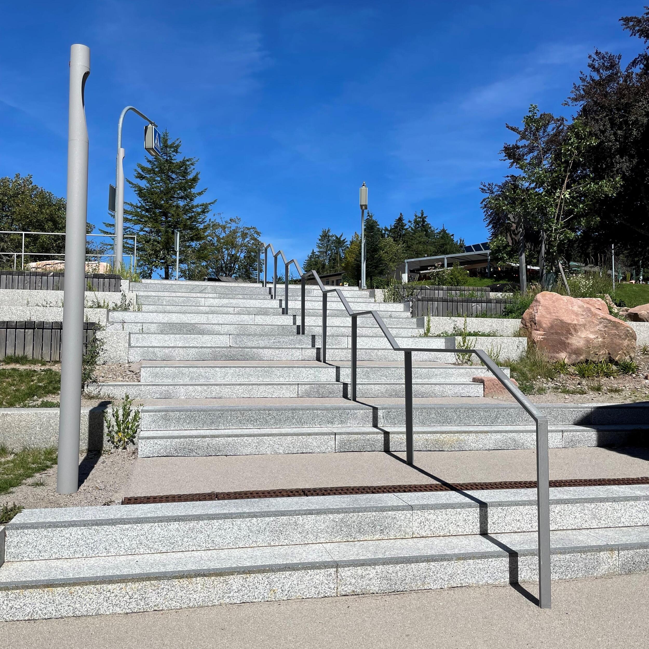 Breite Treppenstufen (Beton mit Granitplatten), Handlauf mittig. Die 15 Stufen sind unterbrochen durch kurze Plateaus.