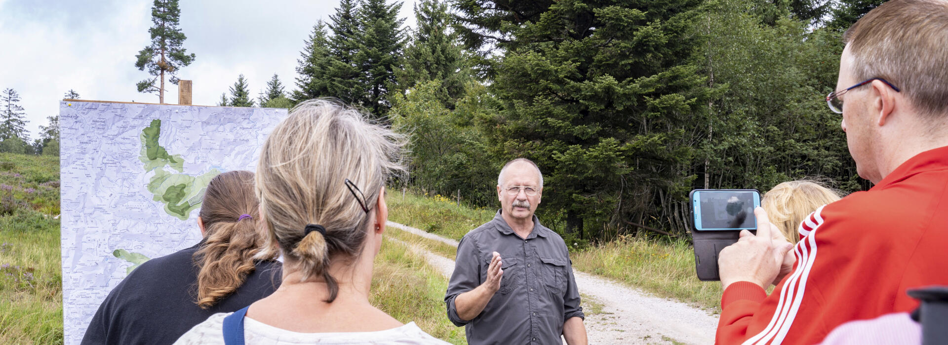 Ein Nationalparkmitarbeiter erklärt einer kleinen Gruppe etwas. Neben ihm hängt eine Karte des Nationalparks an einem Holzgestell. Die Veranstaltung findet im Freien statt.