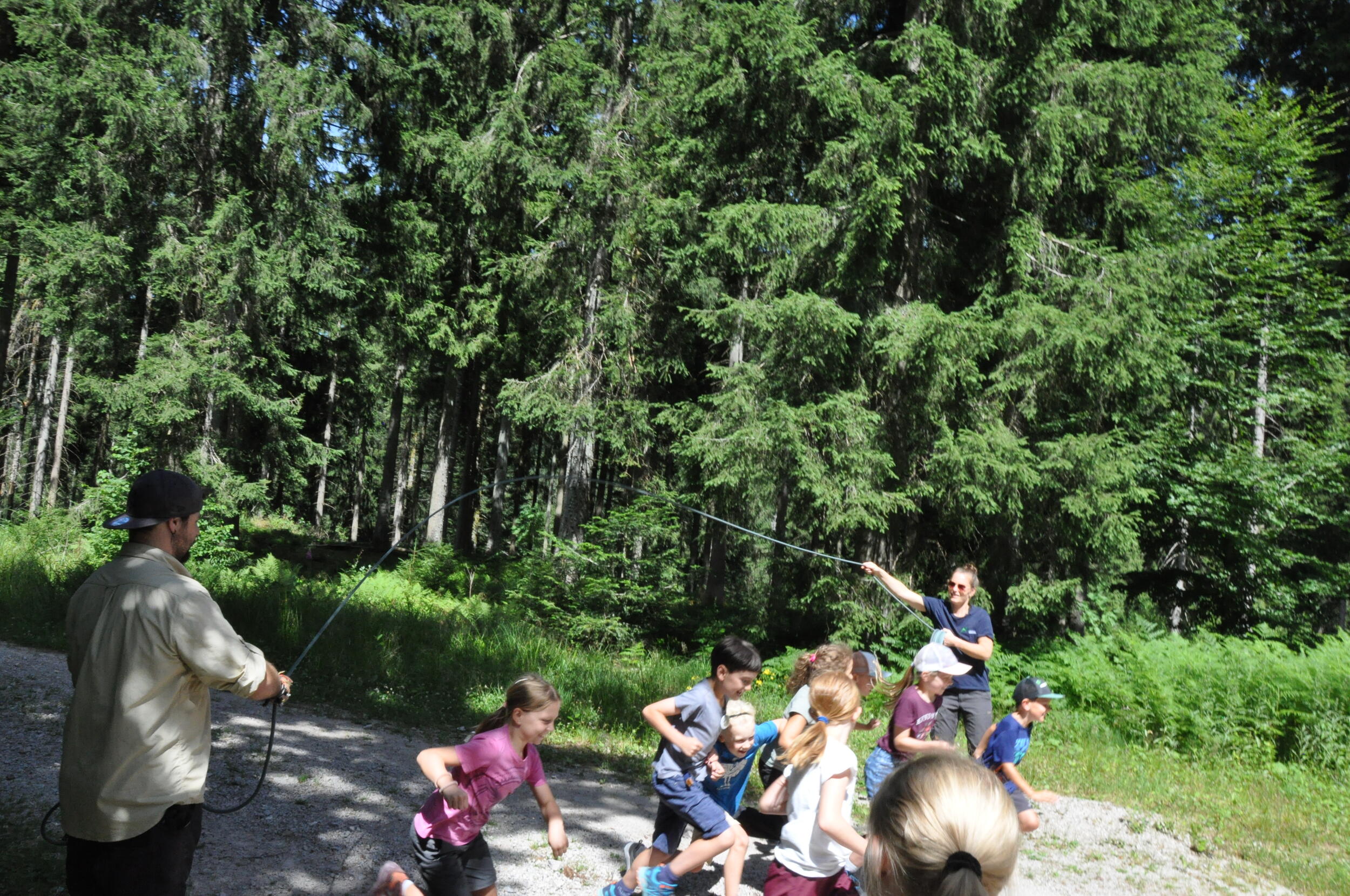 Im Wald auf einem Weg. Zwei Erwachsene schwingen ein langes Seil, unter dem eine Kindergruppe ungestreift durchrennt.