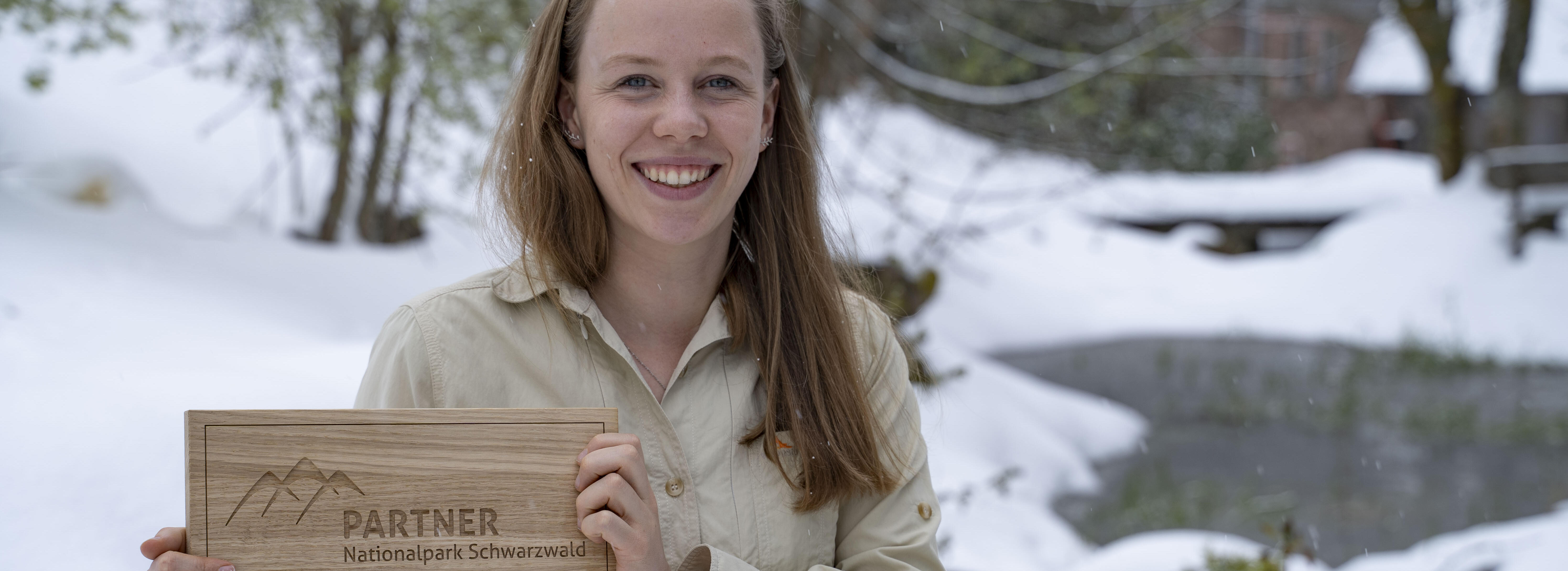 Eine Frau in einem beigefarbenen Hemd steht in einer verschneiten Landschaft und hält ein Holzschild mit der Aufschrift Nationalpark Schwarzwald. Foto: Daniel Müller