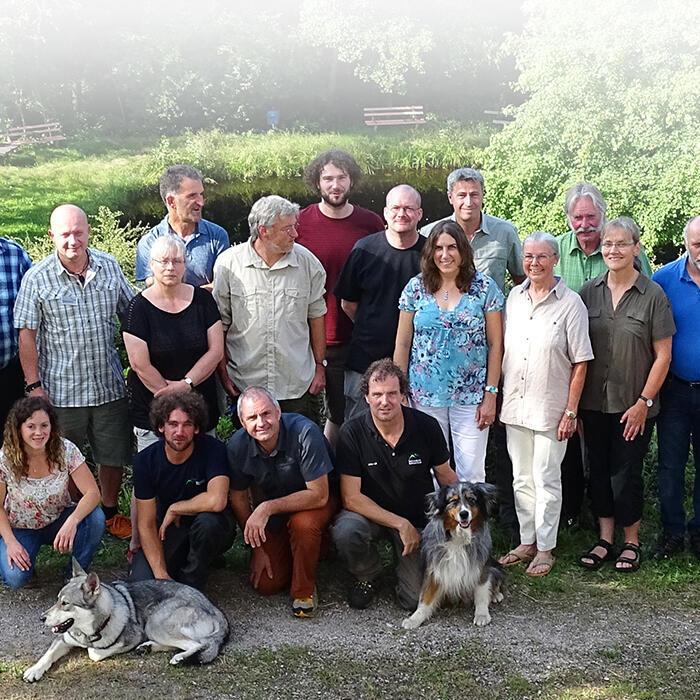 Größere Gruppe Ehrenamtlicher Rangerinnen und Ranger in sommerlicher Kleidung mit See und Bäumen im Hintergrund.