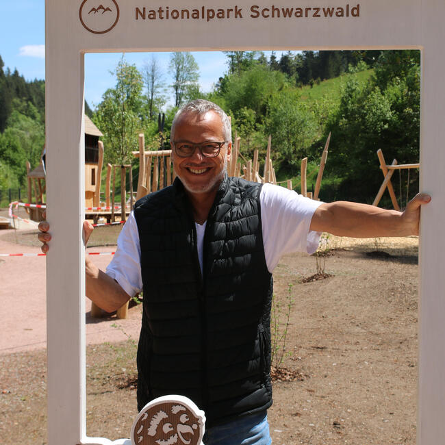 Ein Mann in blauer Weste und weißem Tshirt schaut durch einen weißen Holzrahmen, im Hintergrund ein Spielplatz. Foto: Anne Kobarg