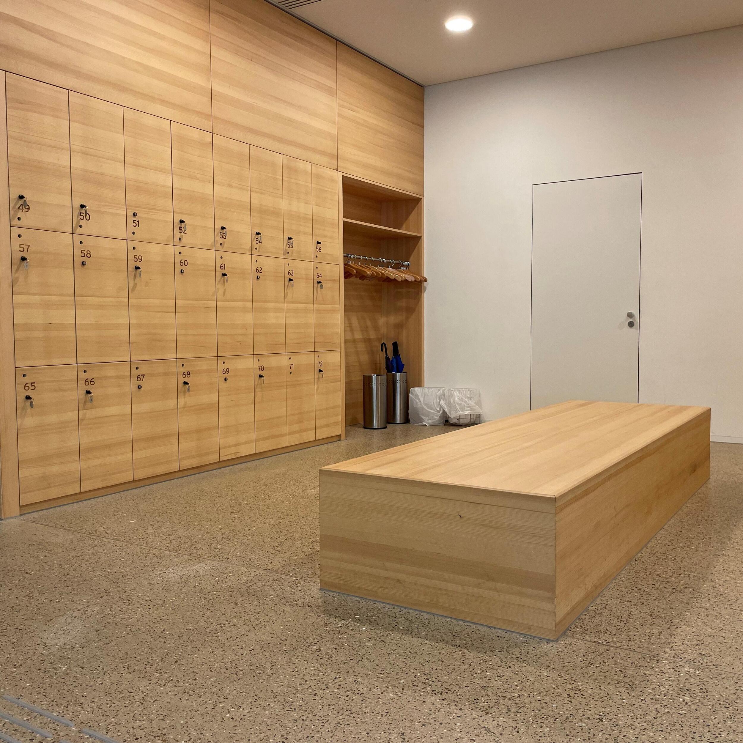 Holzwand mit drei Reihen eingebauter Schließfächer (jeweils ca. 50x30). Rechts in der Wand offene Garderobe mit Kleiderbügeln und Schirmständern. Mit Abstand von 1,50 dazu ein Sitzblock aus hellem Holz. 