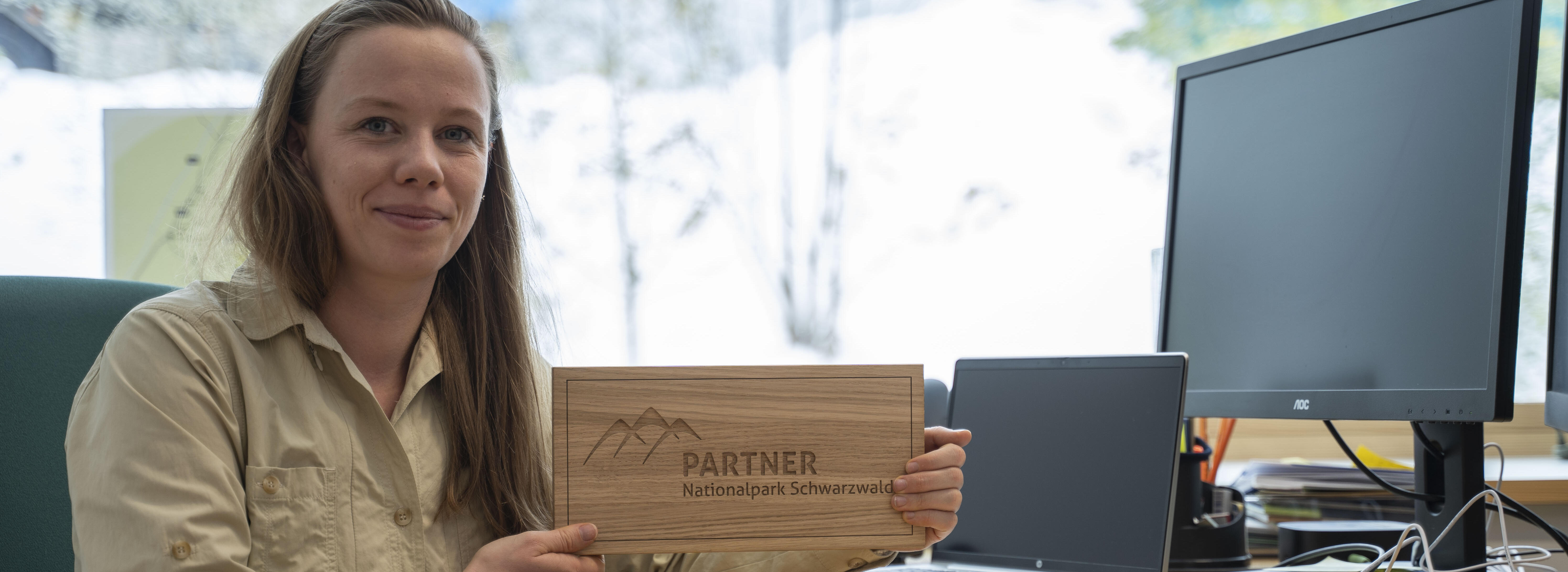 Eine Frau in Nationalparkbluse sitzt am Schreibtisch und hält eine Holzplakette mit dem Schriftzug Partner. Foto: Daniel Müller