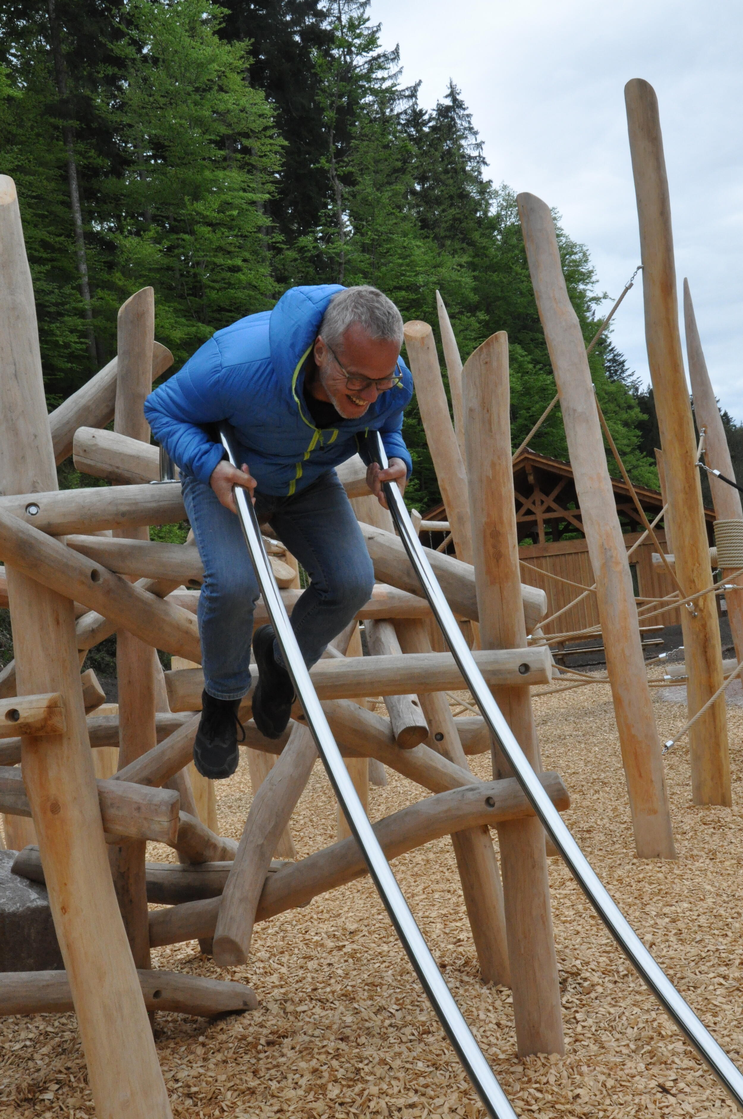 Ein Mann in blauer Jacke ist auf einem Spielplatz mit viel Holz. Er testet eine Kinderrutsche und lacht. 
