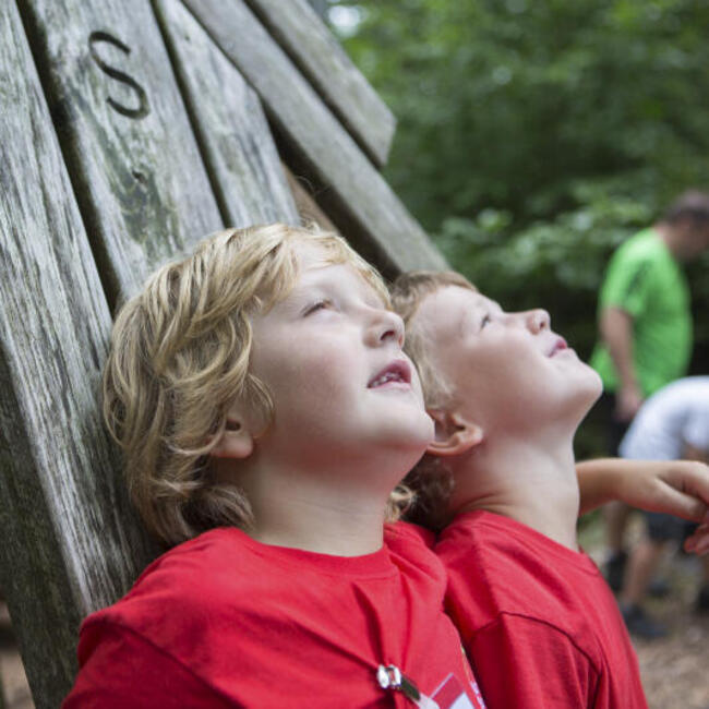 Zwei Kinder in roten T-shirts lehnen an Holzplanken und schauen nach oben. Foto: qu-int.gmbh