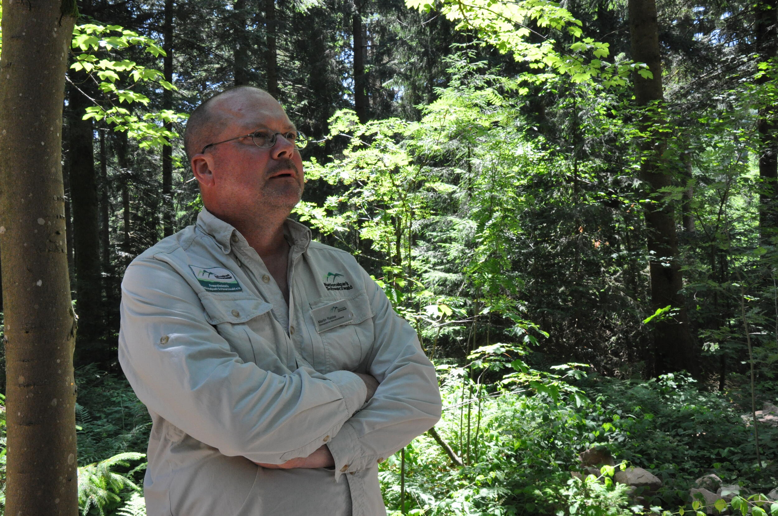 Ein Mann im beigen Hemd und einer Brille steht vor Hintergrund eines Waldes. Seine Arme sind gekreuzt. Er schaut in die Ferne. Foto: Nina Blazon 
