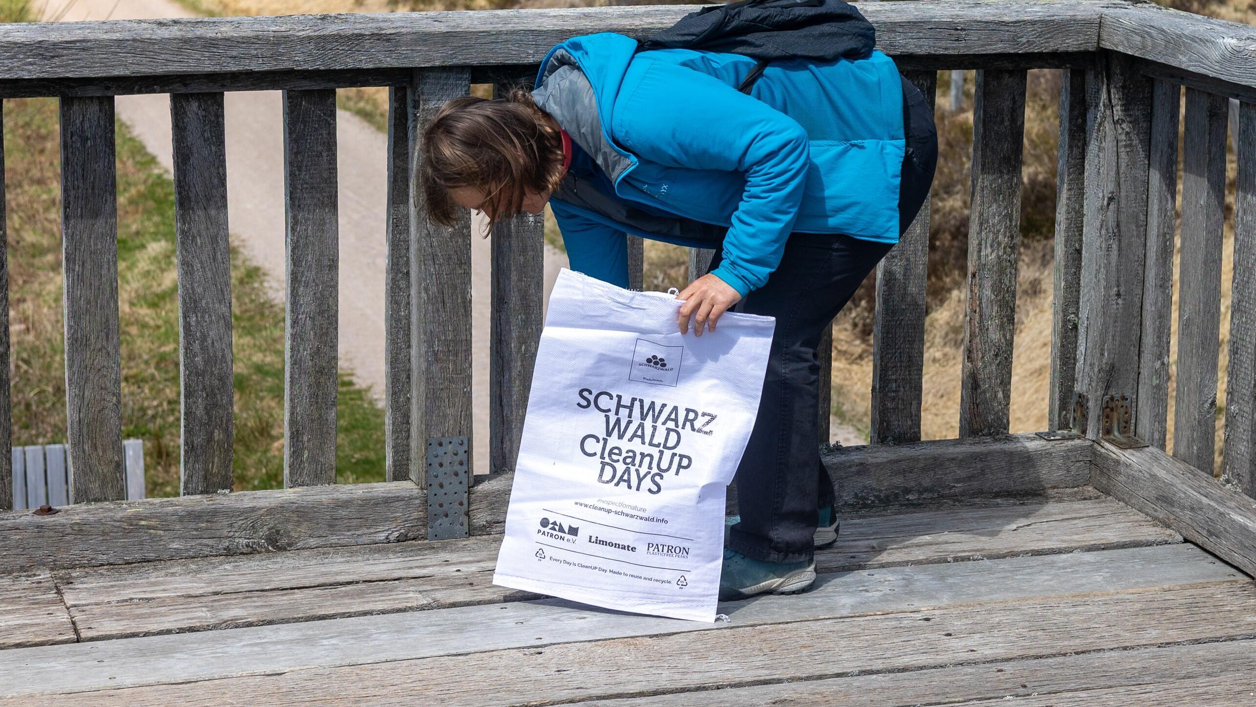 Frau in Wanderkleidung auf einer Holz-Aussichtsplattform. Sie hat eine Tüte in der Hand mit dem Aufdruck "Schwarzwald Cleanup Days" und bückt sich gerade, vermutlich, um Müll aufzusammeln.