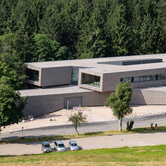 Ein großes Gebäude, bestehend aus mehreren langen, rechteckigen Elementen. Das Gebäude ist holzverschalt und liegt an einer Straße vor einer Nadelwaldkulisse.