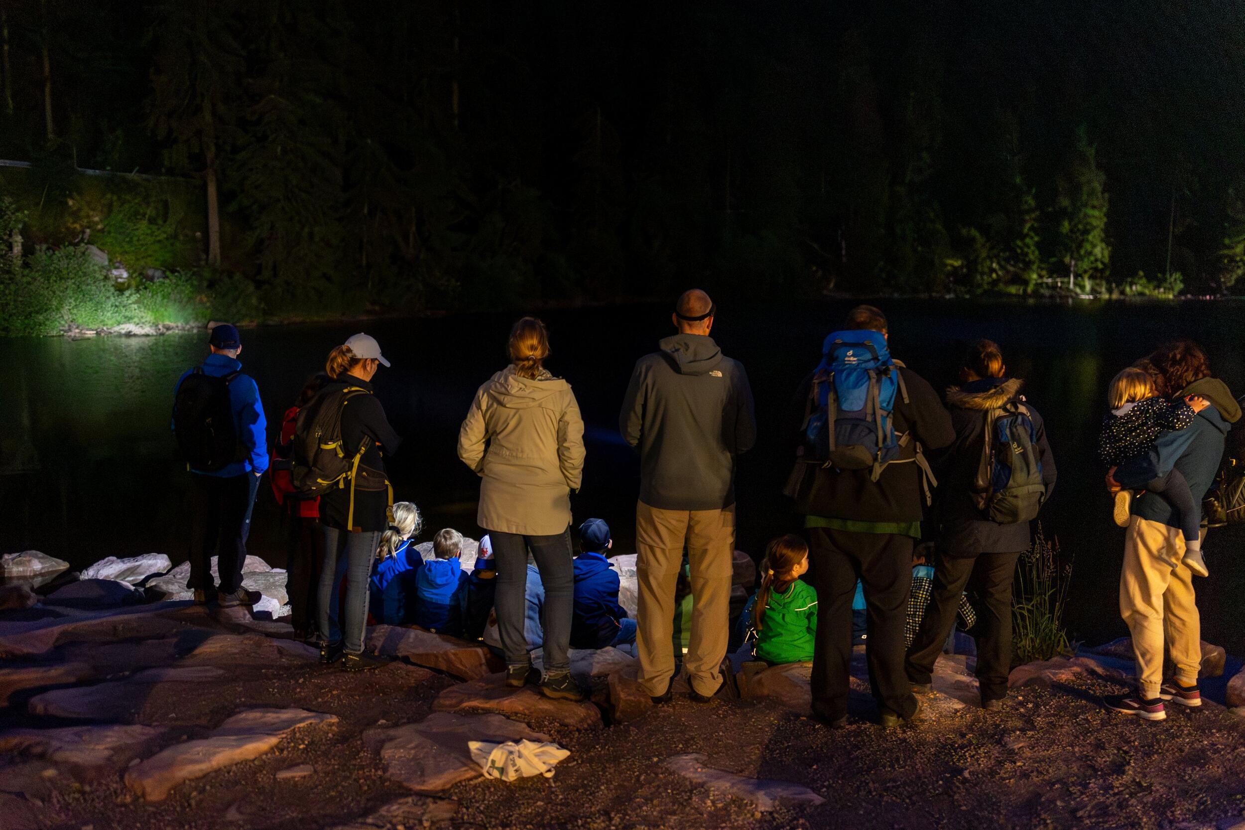 In der Dunkelheit steht und sitzt eine Gruppe Menschen an einem Seeufer.