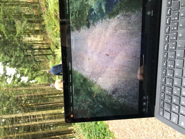 Abbildung 1: Zu sehen ist ein Bildschirm eines Laptops, der von einer Person gehalten wird, die auf einem Wanderweg läuft. Auf dem Bildschirm sind zwei Wanderer von hinten zu sehen.