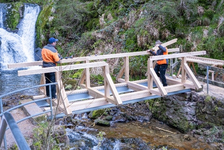 Zwei Männer sind fast fertig mit dem Bau einer Holzbrücke - im Hintergrund ein Wasserfall. Foto: Andreas Forch