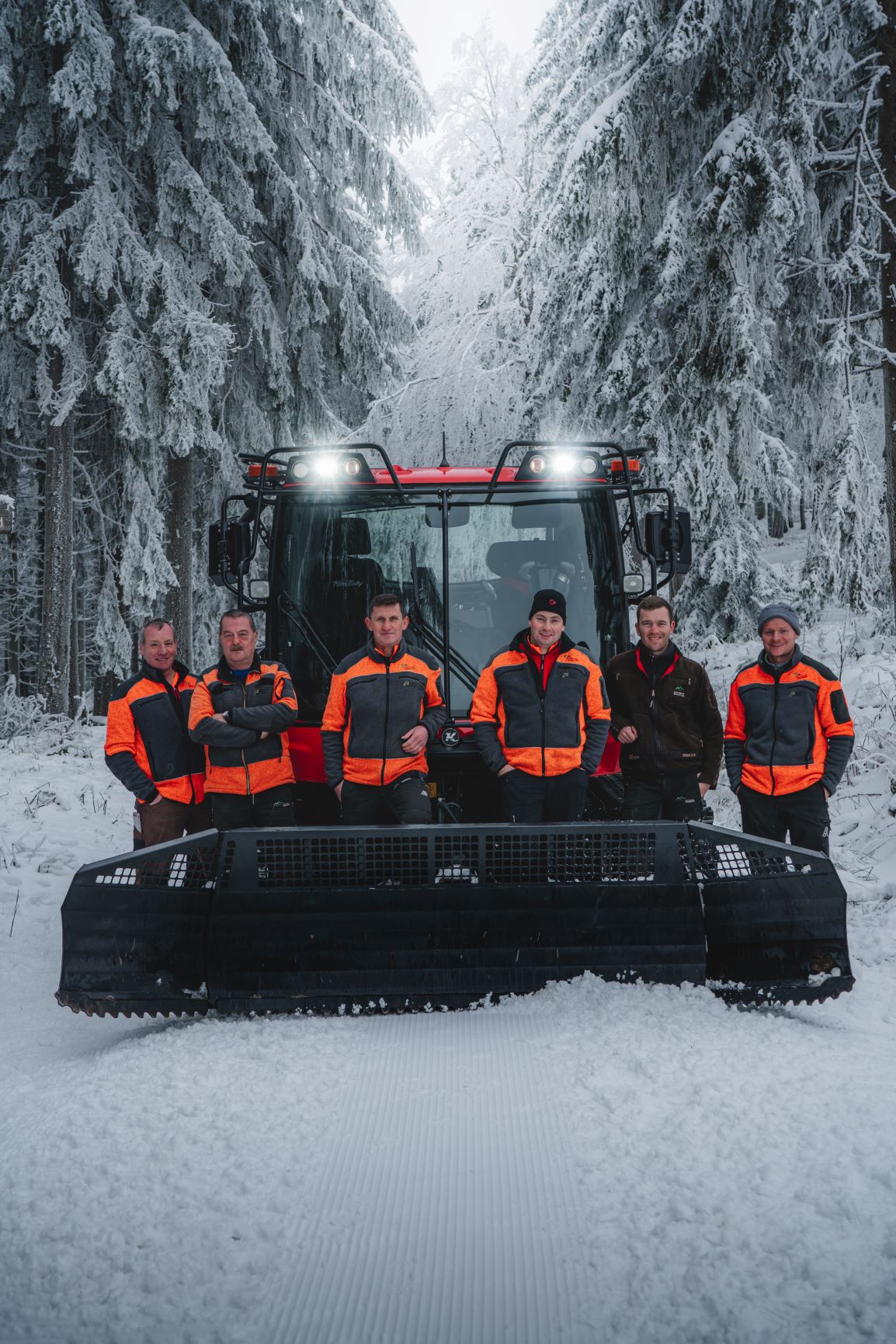 Sechs Männer in orangen Jacken stehen vor einem Schneefahrzeug mitten im Wald. Sie räumen die Loipen. Foto: Tim Tschöpe 