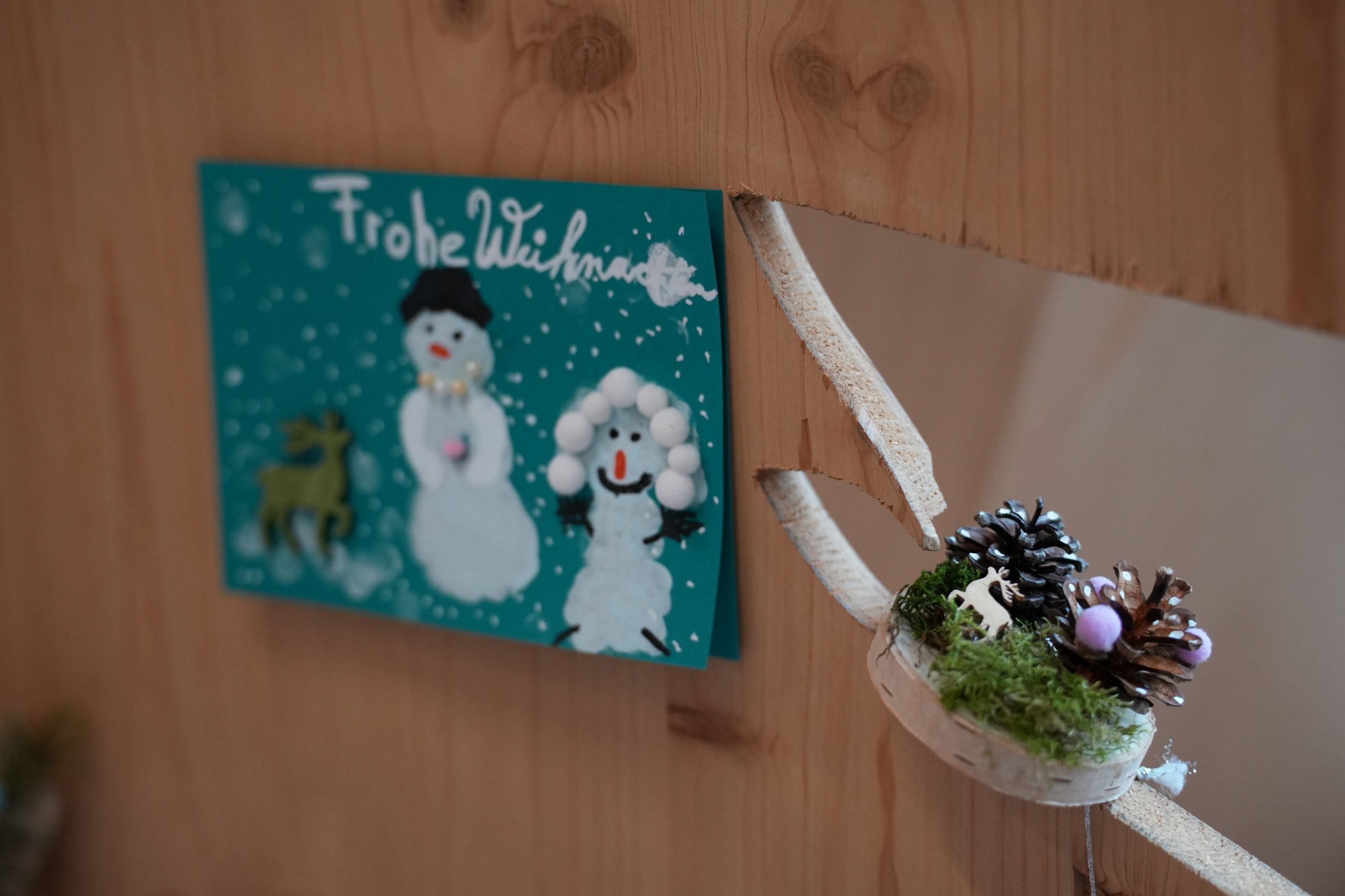 Eine türkise Postkarte mit zwei Schneemännern hängt an einer Holzwand. Daneben eine Birkenscheibe mit Kiefernzapfen. Foto: Ekaterina Nastah