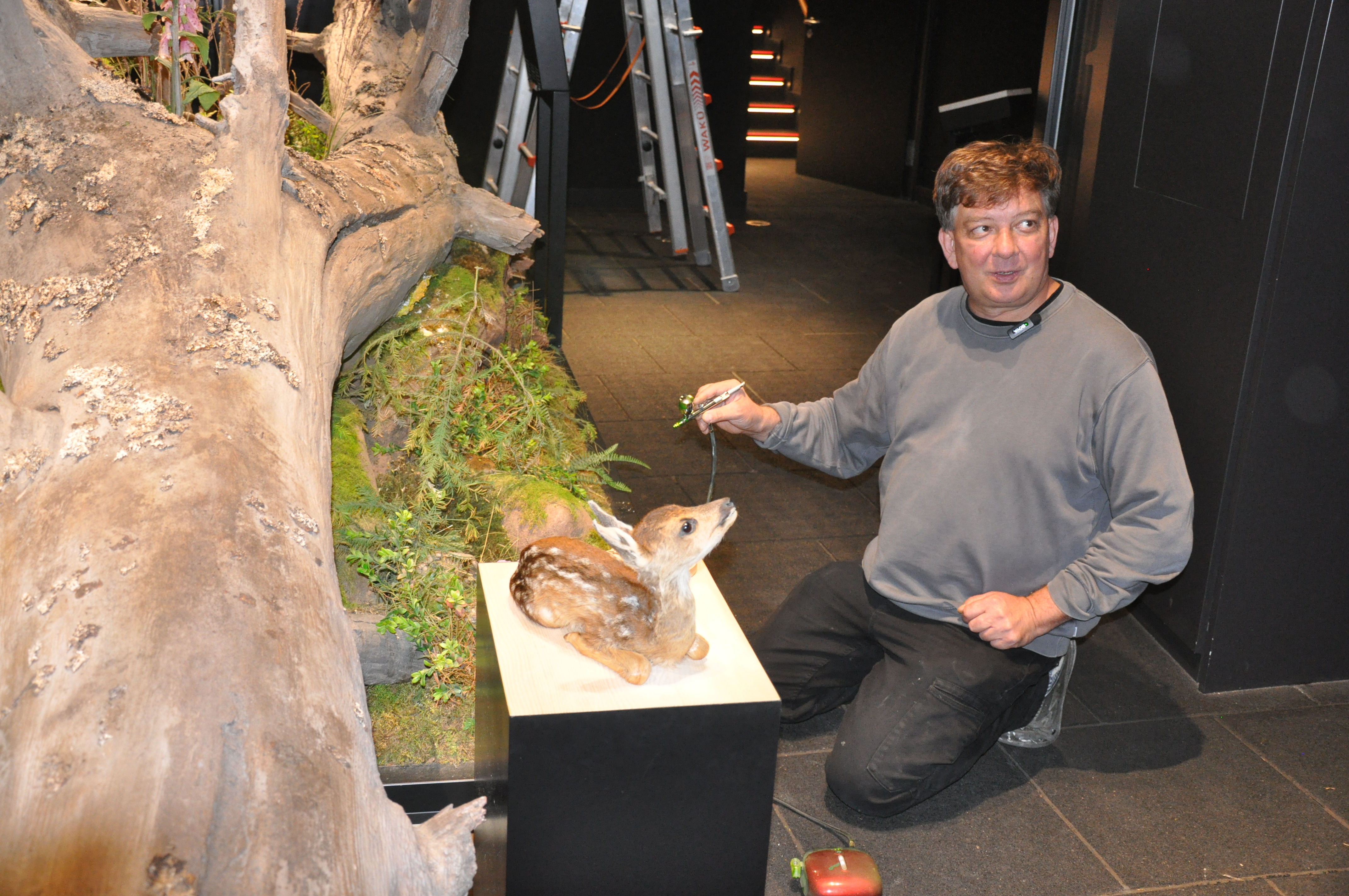 Ein Mann sitzt auf dem Boden neben einem künstlichen Rehkitz auf einem Podest. Er hält ein Instrument mit Farbe in der Hand. Foto: Iris Lemanczyk