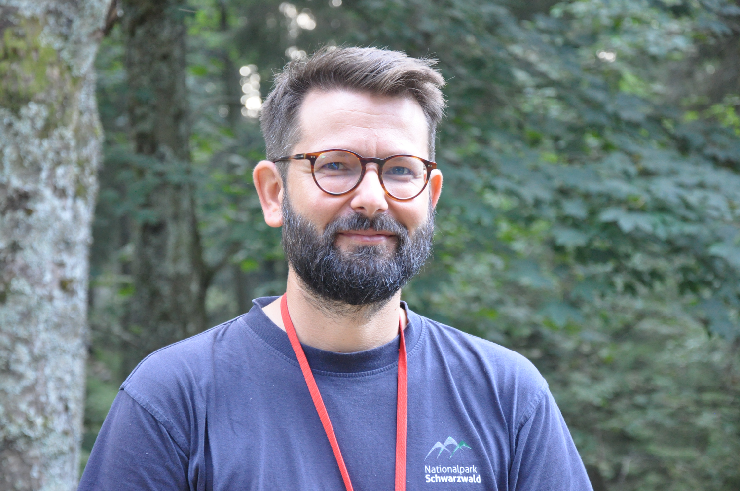 Ein Mann mit Brille und einem roten Band um den Hals trägt ein blaues Tshirt und steht in einem Wald. Foto: Nina Blazon