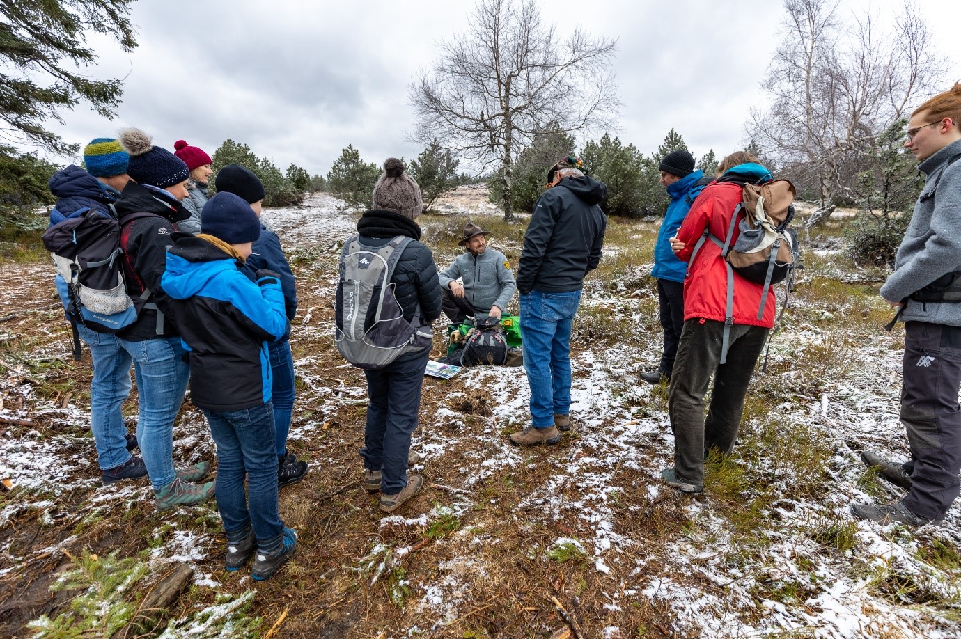 Ein Ranger hockt mit einem Rucksack vor sich auf einem leicht verschneiten Boden. Um ihn herum stehen im Halbkreis Menschen, mit dem Rücken zur Kamera. Foto: Andreas Forch