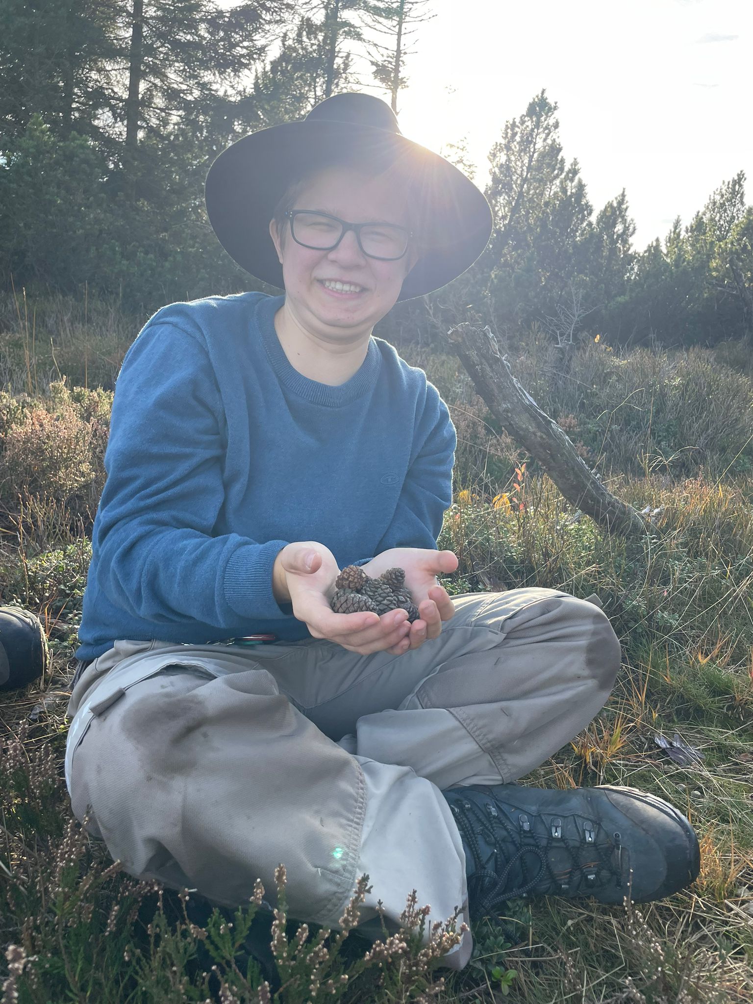 Man sieht eine junge Frau, die einen schwarzen Hut trägt, in der Sonne sitzt und ihn ihren Händen Zapfen hält. Foto: Privat