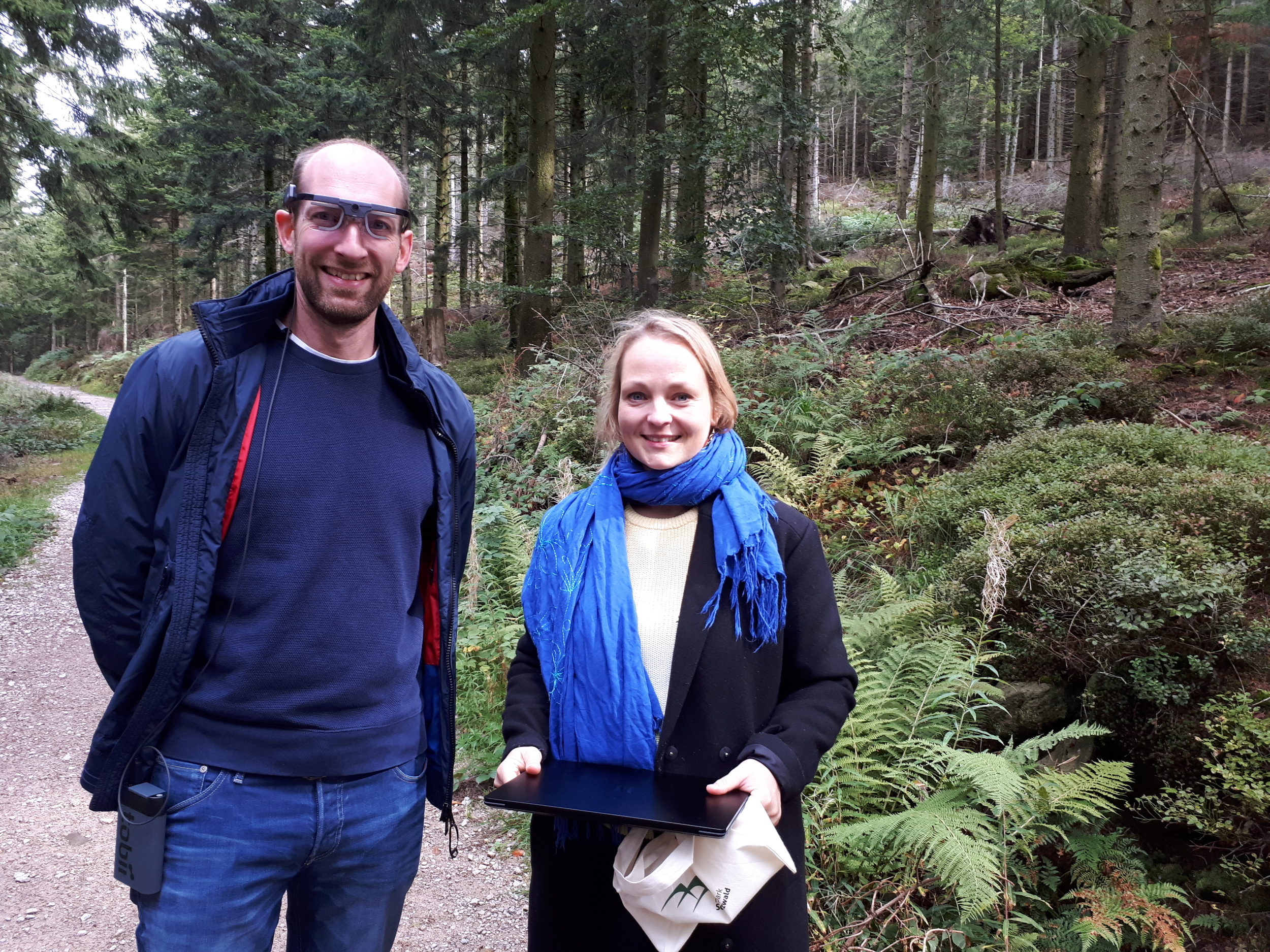Markus Schönberger schulte das Team im Schwarzwald im Umgang mit den Eyetracking-Brillen. Neben ihm: Kerstin Botsch mit dem Laptop zum Verfolgen der Aufnahme. © Susanne Berzborn Ein Mann mit spezieller Brille auf, eine Frau mit einem zugeklappten Laptop in der Hand. Beide stehen nebeneinander auf einem Weg im Wald. Foto: Susanne Berzborn