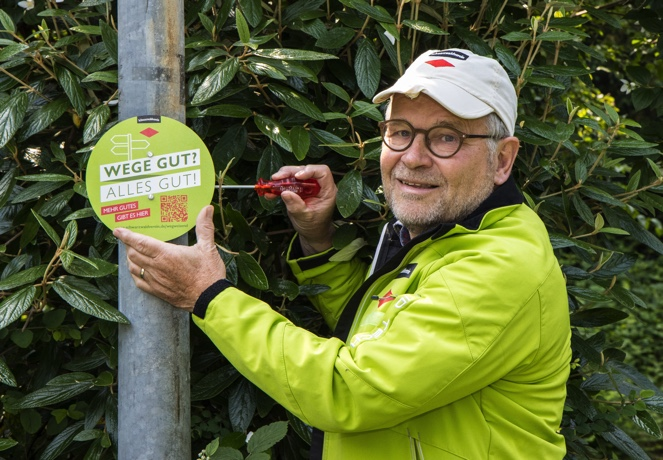Ein Mann mit hellgrüner Jacke und heller Schirmmütze montiert ein grünes Schild mit der Aufschrift "Wege gut - alles gut" an einer Stange. Foto: Schwarzwaldverein