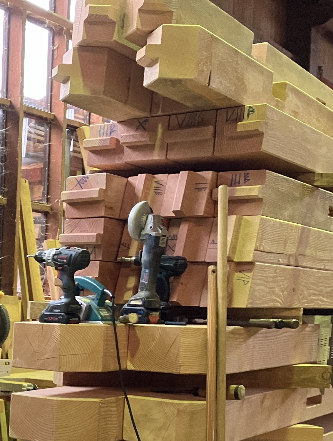 Für mich sind die Zeichen Gekritzel, für Stefan ist es der Plan, wie die Hütte zusammengebaut werden muss. © Nina Blazon Ein Stapel mit vielen Holzbalken, auf denen Zeichen zu sehen sind. Davor stehen ein Akkuschrauber und eine Handsäge. Foto: Nina Blazon