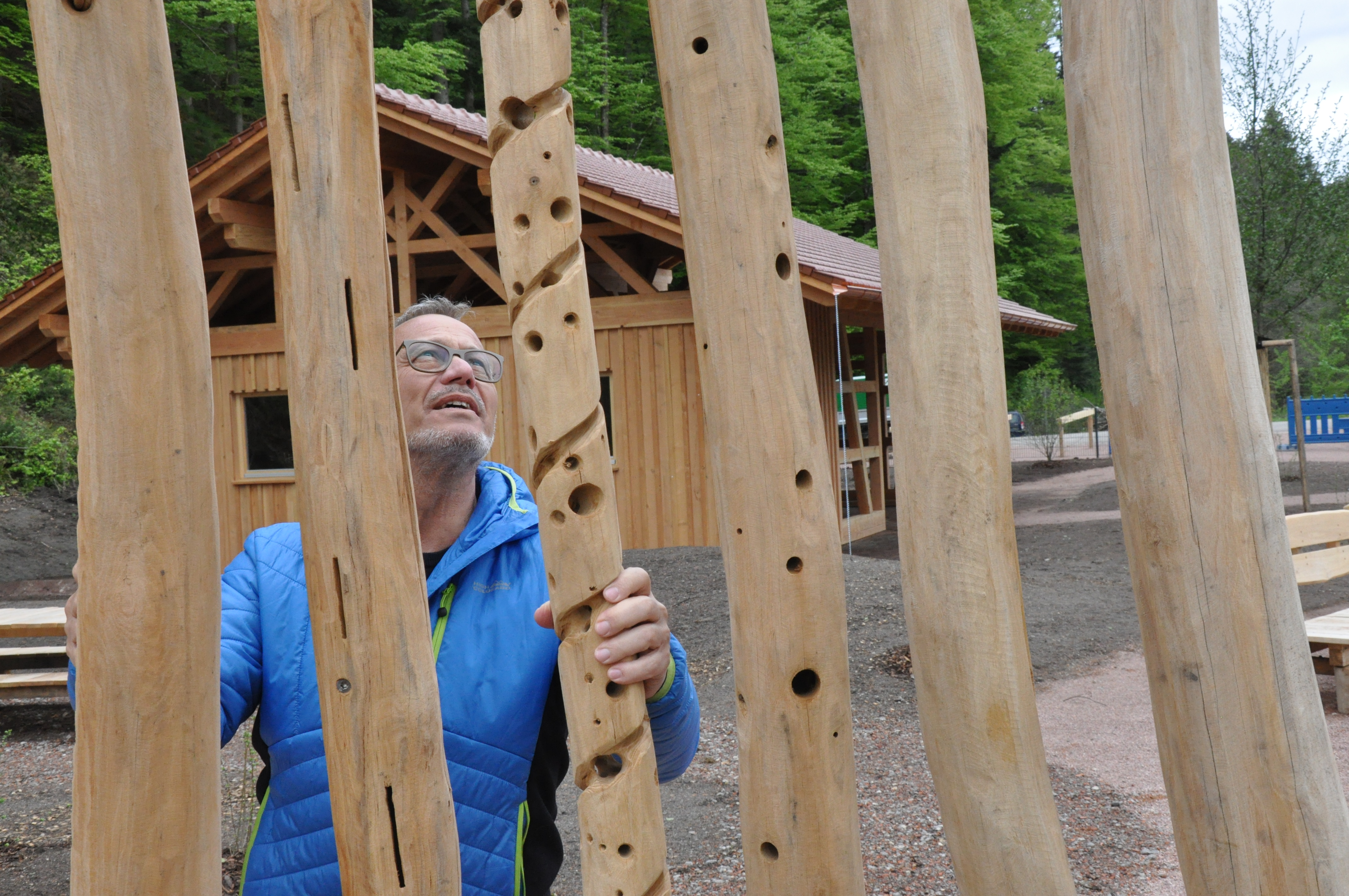 Ein Mann in blauer Jacke steht vor hölzernen Balken. Hinter ihm ist eine Hütte. 