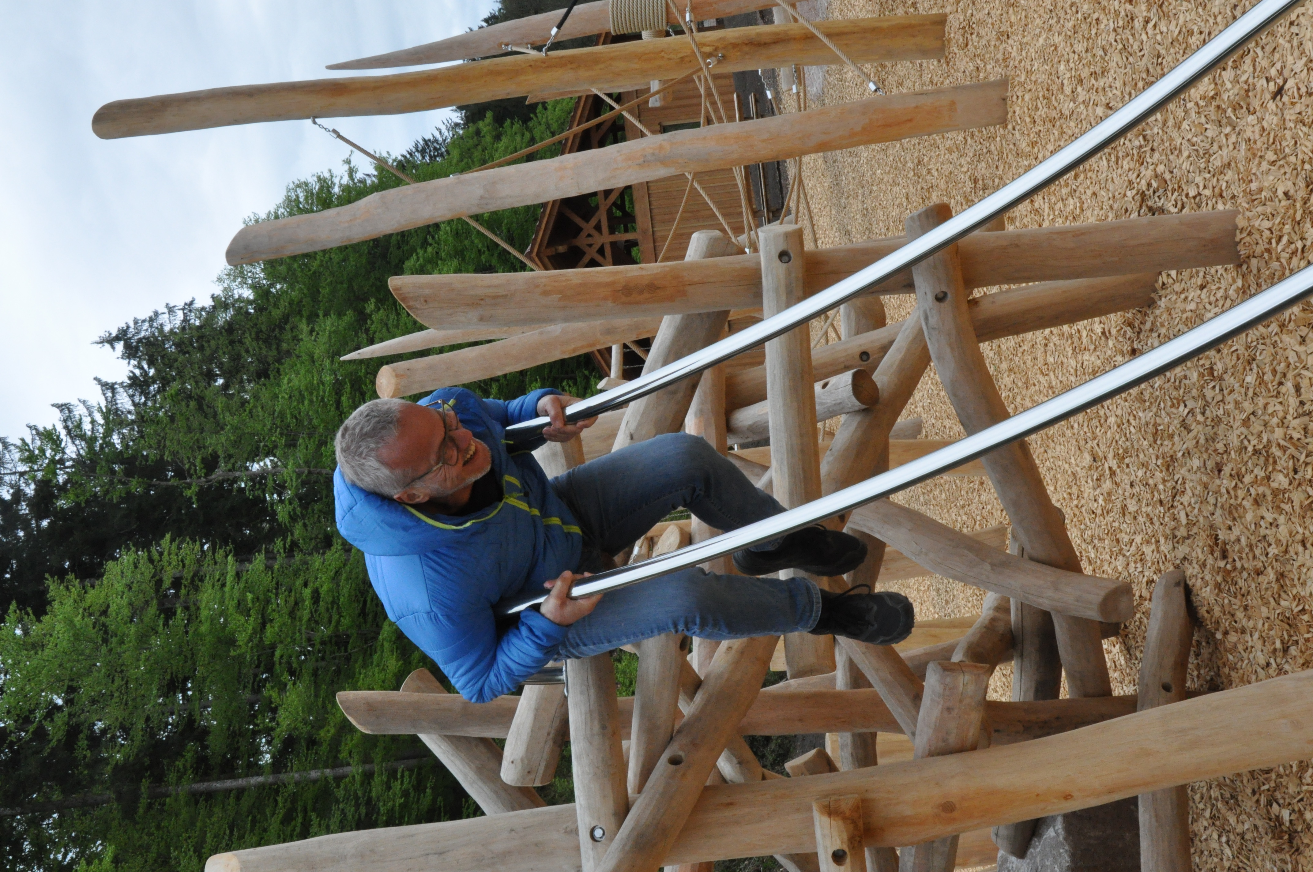 Ein Mann in blauer Jacke ist auf einem Spielplatz mit viel Holz. Er testet eine Kinderrutsche und lacht. 