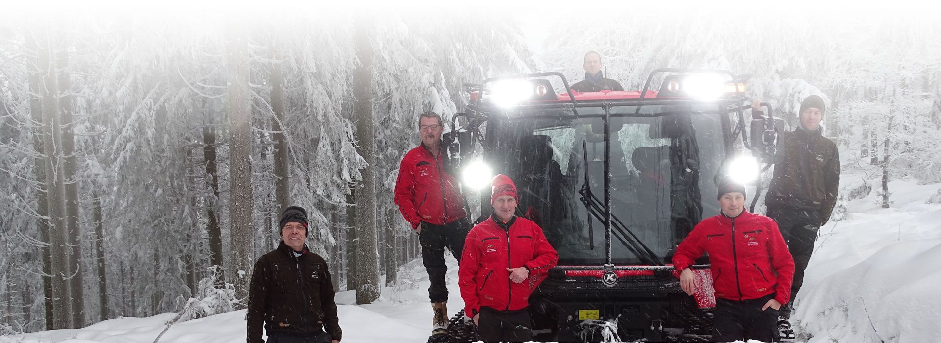 Mehrere Männer in roten und braunen Nationalparkjacken haben sich vor einem verschneiten Winterwald um einen roten Pistenbully gruppiert. Foto: Friederike Scharfe