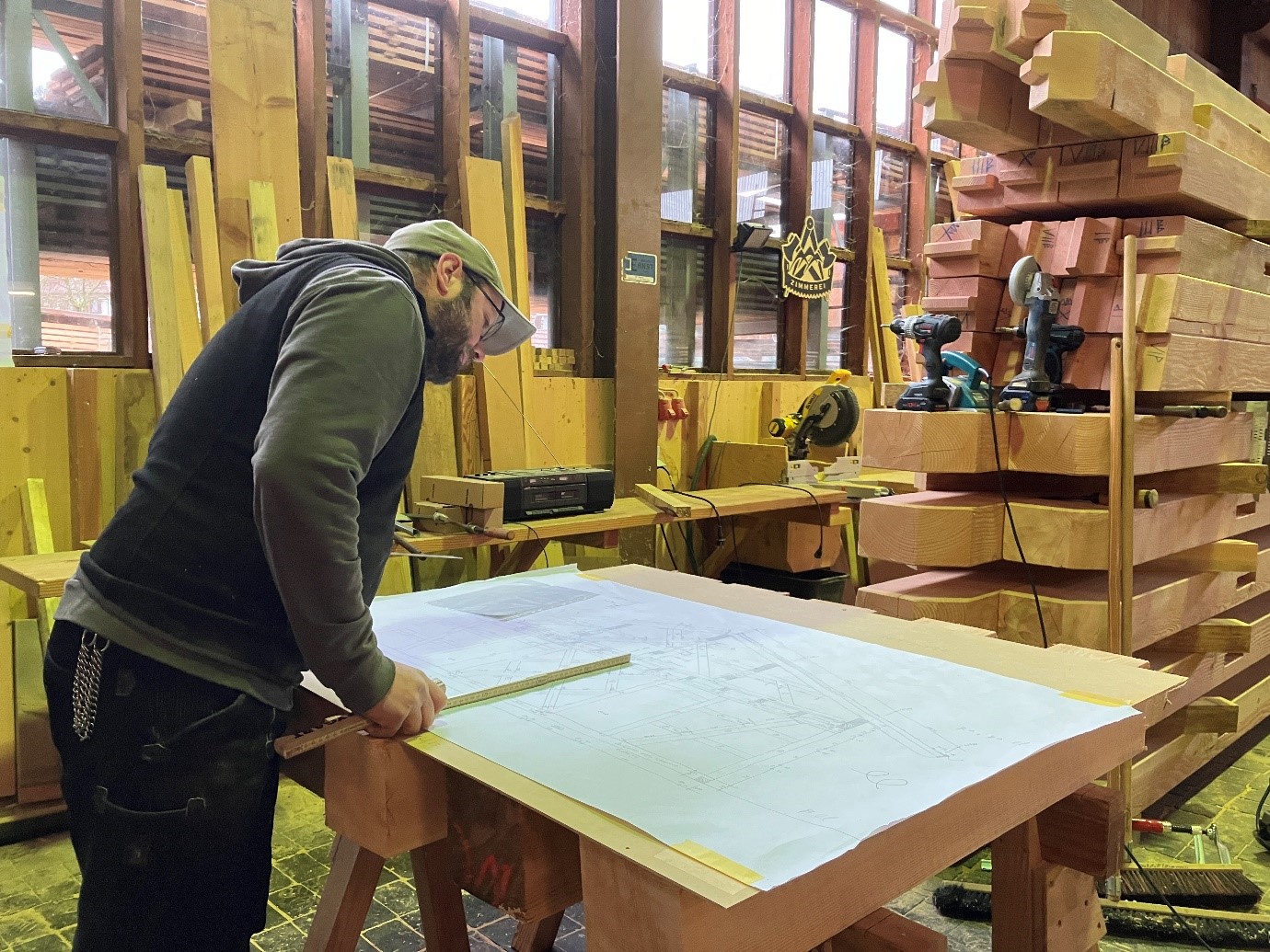 Auf Stefans Rechenkünste kommt es an. © Nina Blazon Ein Mann steht an einem Tisch, in der rechten Hand hält er einen Zollstock. Auf dem Tisch liegt ein Plan, den er konzentriert betrachtet. Im Hintergrund ein Stapel mit kantigen Hölzern. Foto: Nina Blazon
