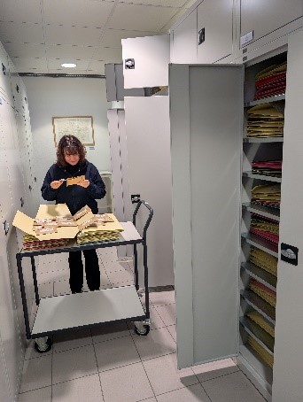 Eine schwarz gekleidete Frau mit dunklen Haaren steht in einem Raum mit vielen grauen Schränken, zum Teil geöffnet. Sie steht hinter einem Rollwagen, auf dem Naturmaterial liegt und betrachtet etwas in ihren Händen. Foto: Naturkundemuseum