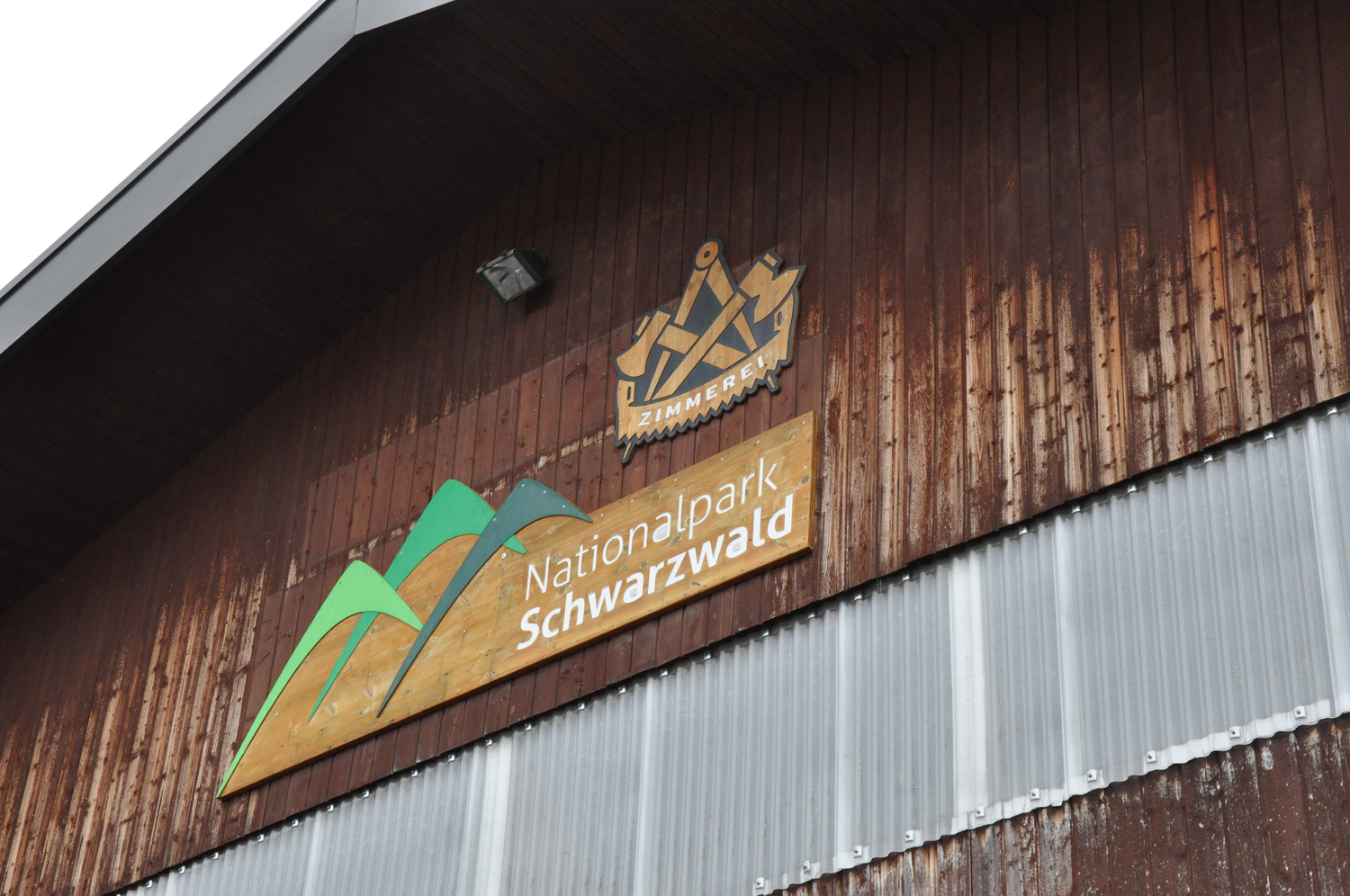 Seit 2019 gehört die Zimmerei zum Nationalpark. © Nina Blazon An einem Holzhaus hängen unter dem Dachgiebel zwei Schilder, auf dem einen steht Zimmerei, auf dem unteren Nationalpark Schwarzwald. Foto: Nina Blazon