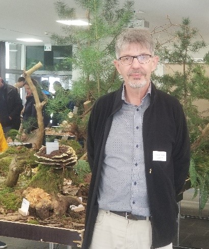 Ein Mann in schwarzer, offener Jacke und grauem Hemd steht vor einem Glaskasten mit Naturmaterialien im Inneren. Foto: Naturkundemuseum