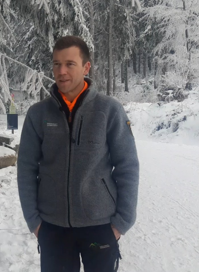 Ein Mann in grauer Jacke "Nationalpark Schwarzwald" steht vor winterlichem Hintergrund. Er hat seine Hände in den Taschen. Er ist Leiter des Loipenteams. Foto: Tim Tschöpe 
