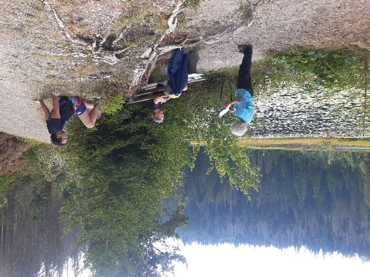 Befragung am Huzenbacher See im Nationalpark Schwarzwald. © Mareike Garms (Nationalpark Schwarzwald) Eine Frau Blick auf einen Zettel, zwei Menschen sitzen am Boden am Ufer eines Sees und schauen zu ihr. Foto: Mareike Garms