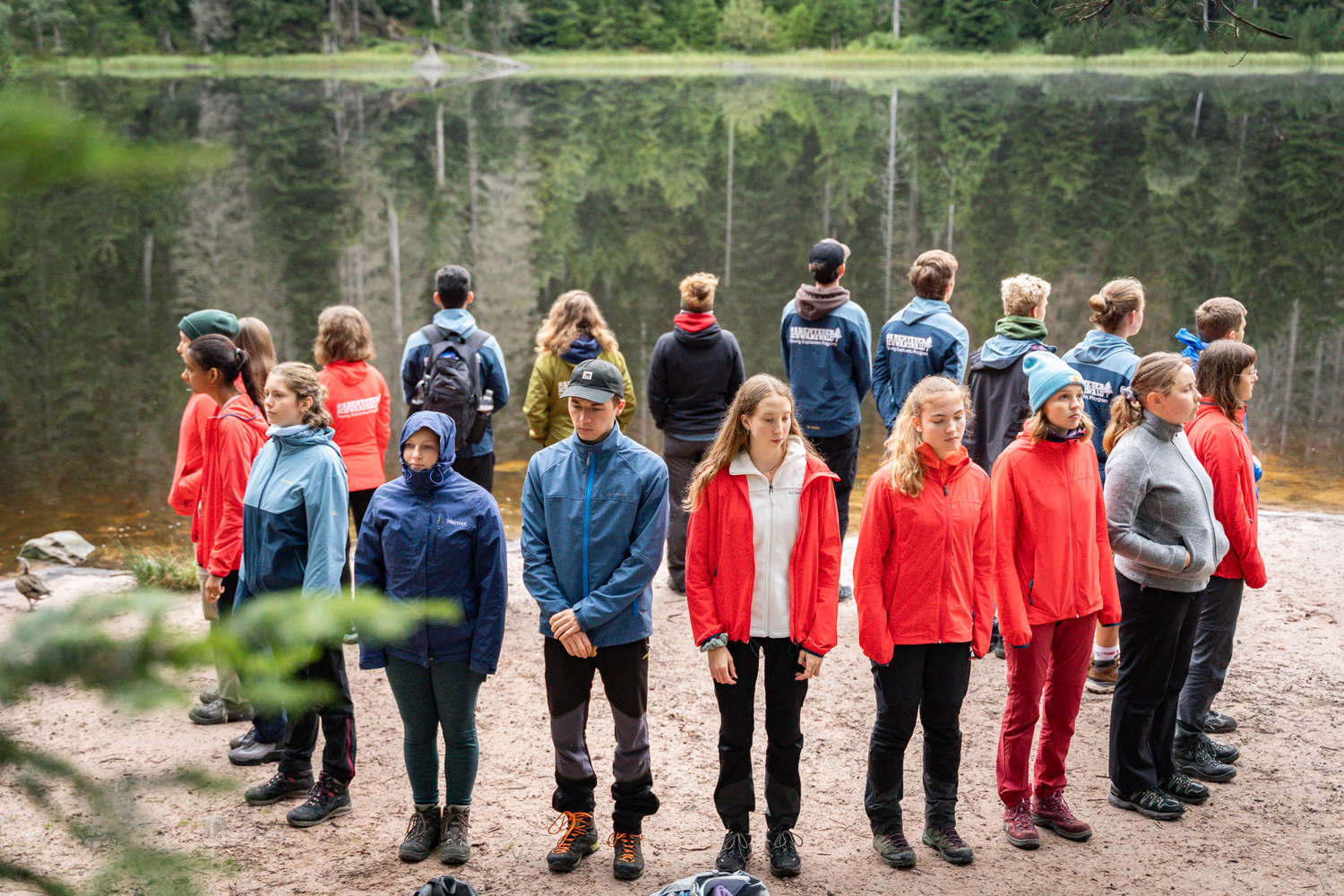 Jugendliche stehe in einem Kreis mit den Rücken zueinander an einem See, in dem sich Bäume spiegeln. Foto: A. Schneider