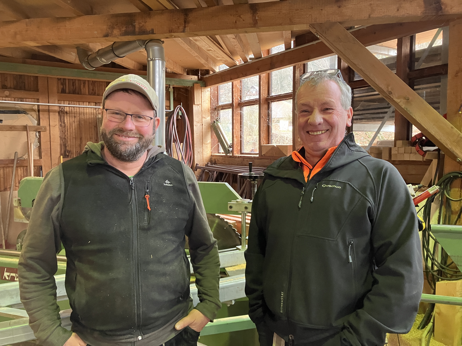 Die Huber-Brüder in der Zimmerei: links Stefan, rechts Markus. © Nina Blazon Zwei Männer lächeln in die Kamera, der linke trägt ein Käppi. Im Hintergrund ist eine Holzwerkstatt zu sehen. Foto: Nina Blazon