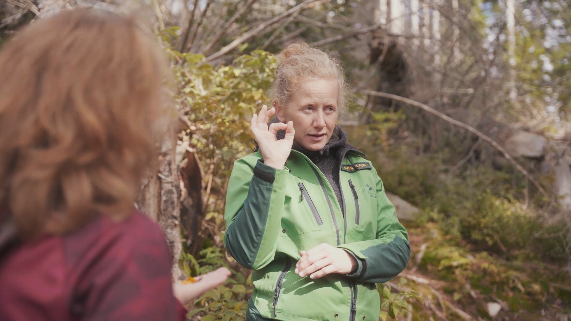 Eine Frau mit hochgestecktem Haar steht in einer grünen Outdoor-Jacke im Wald und nutzt Gebärdensprache im Gespräch mit einer anderen, unscharf im Vordergrund stehenden Frau. Foto: Juliane Schrempp 