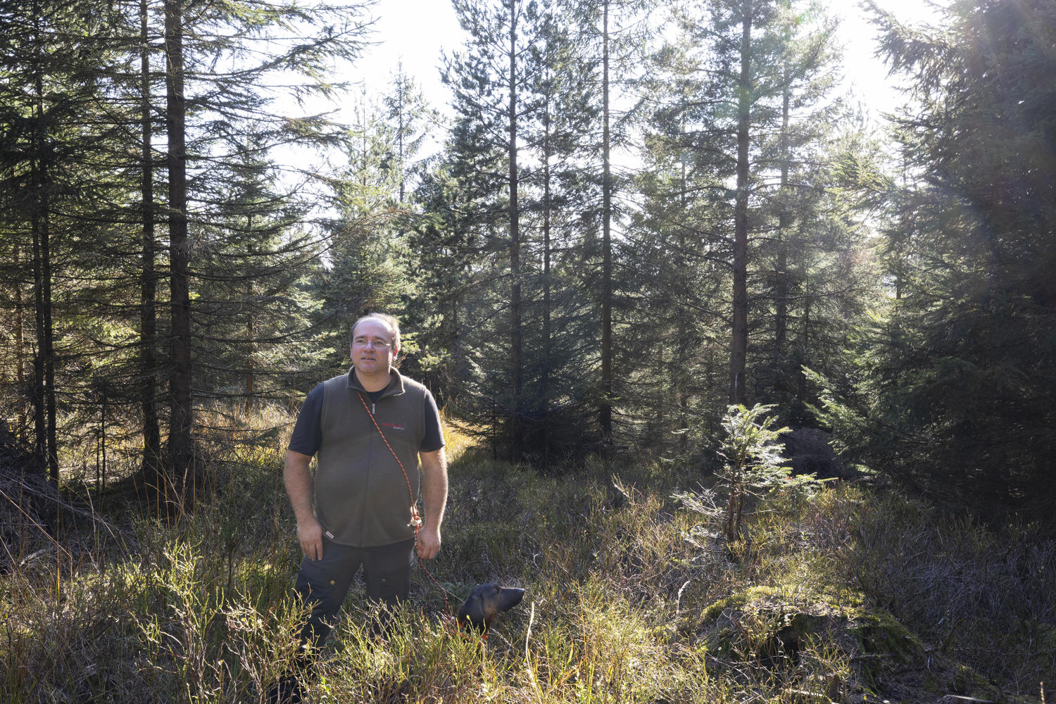 Ein Mann im Tshirt und mit olivegrüner Weste steht auf einer sonnenbeschienenen Waldlichtung. Links neben ihm steht ein Hund. Foto: Ulrike Klumpp