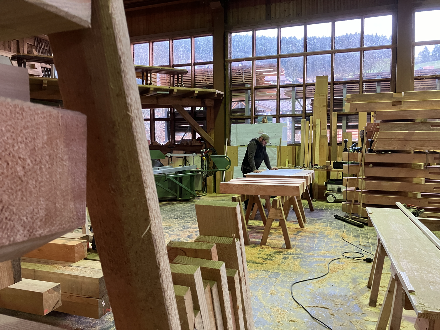 In der Zimmerei dreht sich momentan alles um die Gartenschauhütte. © Nina Blazon Blick in eine Halle mit viel Holz, im Hintergrund arbeitet ein Mann an einem Holztisch. Ganz hinten große Fenster, die in kleine Quadrate unterteilt sind. Foto: Nina Blazon